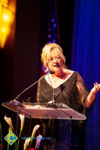 Woman wearing a black evening dress speaking on stage at the podium.