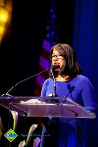 Woman with short dark hair and glasses wearing a blue dress speaking at the podium.