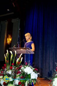 A woman with short blond hair and wearing a blue dress, speaking at the podium.