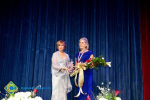 President, JoAnna Schilling, holding an award and standing with a woman in a blue dress who is holding a bouquet of flowers.