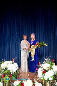 President, JoAnna Schilling, holding an award and standing with a woman in a blue dress who is holding a bouquet of flowers.