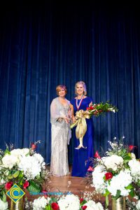 President, JoAnna Schilling, holding an award and standing with a woman in a blue dress who is holding a bouquet of flowers.