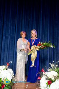 President, JoAnna Schilling, holding an award and standing with a woman in a blue dress who is holding a bouquet of flowers.