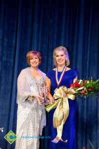 President, JoAnna Schilling, holding an award and standing with a woman in a blue dress who is holding a bouquet of flowers.