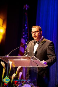 A man in a black suit and tie speaking at the podium.