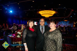 Lynette Young with man in suit, grey shirt and tie and a woman with long brown hair, black dress and red scarf around her neck.