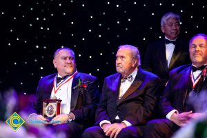 Four men in black suits and tie sitting on stage,