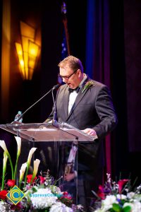 A man in a black suit and tie speaking at the podium.