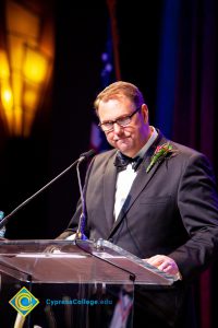 A man in a black suit and tie speaking at the podium.