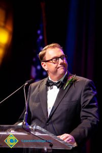 A man in a black suit and tie speaking at the podium.