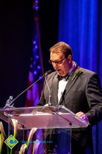 A man in a black suit and tie speaking at the podium.