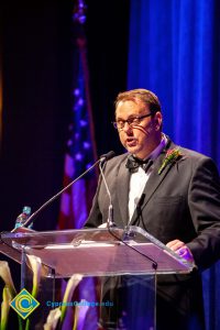 A man in a black suit and tie speaking at the podium.