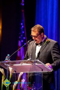 A man in a black suit and tie speaking at the podium.