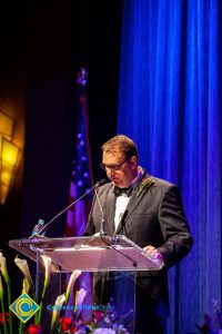 A man in a black suit and tie speaking at the podium.