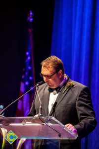 A man in a black suit and tie speaking at the podium.