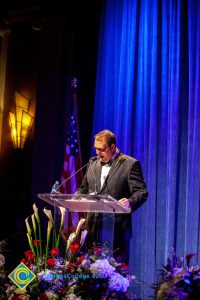 A man in a black suit and tie speaking at the podium.