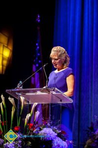 A woman with short blond hair and wearing a blue dress, speaking at the podium.