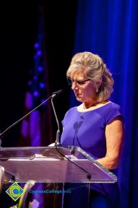 A woman with short blond hair and wearing a blue dress, speaking at the podium.