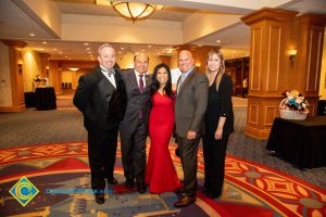 Three men in suits, a woman in a red dress, and a woman in a black pantsuit.