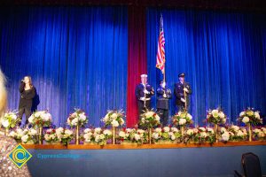 A woman in black on the stage with a microphone and three members of the military holding a flag and rifles.