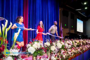 A girl in a blue dress, a girl in a red dress, and a young man in a tan shirt on stage holding a white broom.