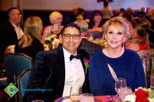 A man in a black jacket, white shirt and bowtie sitting with a woman with blong hair and a blue dress.