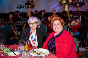 Woman in a black and purple floral blouse with a red jacket and a woman in a black jacket and white shirt smiling,