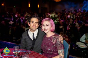 A man with a grey and black sweater, white shirt, and black tie with his arm around a young lady with purple hair and a purple dress.