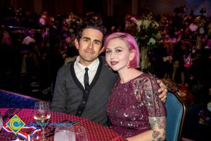 A man with a grey and black sweater, white shirt, and black tie with his arm around a young lady with purple hair and a purple dress.