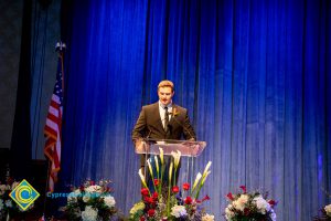 Man in a suit and tie on stage at the podium with a blue curtain in the background and the American flag to the left.