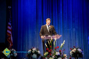 Man in a suit and tie on stage at the podium with a blue curtain in the background and the American flag to the left.