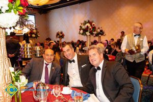 Three men in suits and tie smiling at the dining table with a waiter walking behind them.