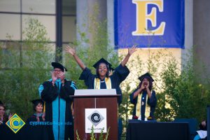 A young lady in graduation regalia at the podium holding her hands in the air while President Bob Simpson applauds and another person cheers in the background.