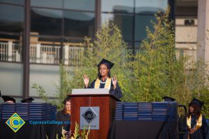 A young lady in graduation regalia at the podium holding her hands up during commencement.