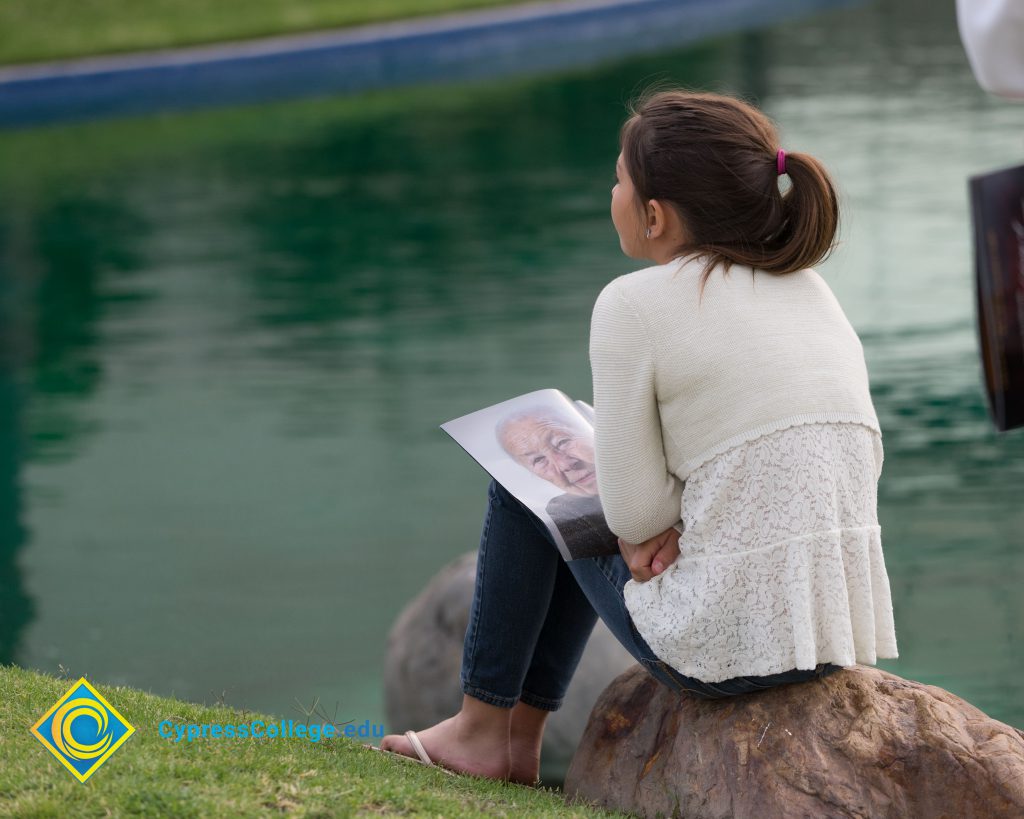 Young female wearing white sweater, jeans, and sandals, sitting on a rock next to the pond with the Yom HaShoah program open in her lap