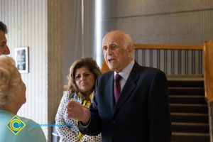 Elderly man wearing dark suit and maroon tie speaking to elderly woman