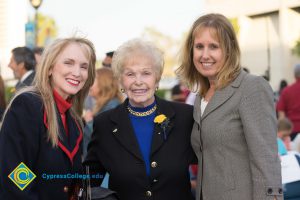 Holocaust survivor wearing black blazer and blue sweater, standing with woman wearing black and red blazer with red sweater and woman wearing tweed blazer and white top