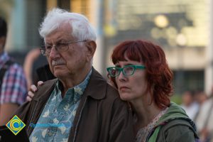 Older man wearing glasses and red-haired woman wearing green glasses watching solemnly from the audience