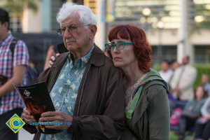 Older man wearing glasses and holding Yom HaShoah program and red-haired woman wearing green glasses watching solemnly from the audience