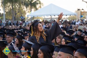 Graduates in regalia sit in the audience at commencement while one stands and raises her hand