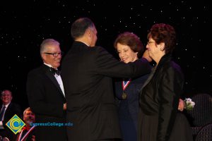 President Bob Simpson and a woman in a black suit look on with a smile as a gentleman in a black suit places a medal around a woman's neck at the 40th Annual Americana Awards.
