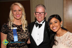 President Bob Simpson smiling beside a blonde woman in black lace dress and a young woman with dark brown hair in a white dress.