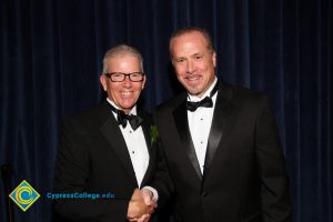 President Bob Simpson and a man with a moustache and beard wearing tuxedos, shaking hands and smiling.