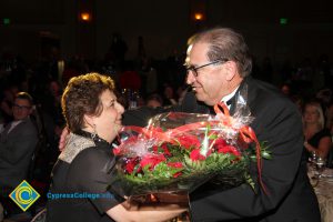 A woman with short brown hair in a black blouse, being presented with a large bouquet of red roses from a gentleman in a black suit.