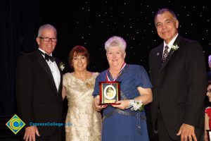 A woman with silver hair and a blue dress holding a award with President Bob Simpson, a woman in a white dress and a man in a dark suit and tie by her side.