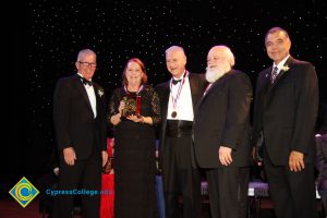 A woman in a black dress holding a award with President Bob Simpson and three men in suits and tie by her side.