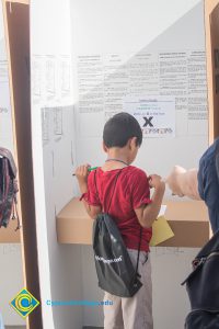 A little boy in a red shirt with a black backpack standing in a cardboard cubicle.