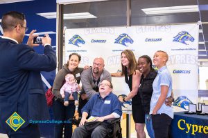 A young man in a wheelchair surrounded by a group of men and women while a man in a suit takes a picture of them.