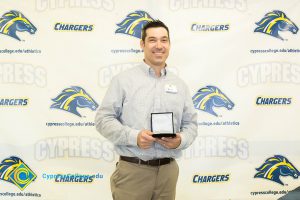 Man in a light blue shirt and tan pants holding a medal.