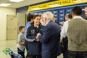 Gentleman with grey hair, glasses and blue suit shaking hands with a smiling young lady in a blue jacket with white striped on the sleeve.